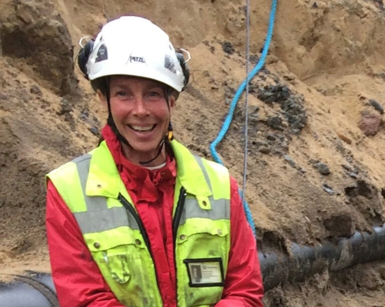 Nainen hymyilee valkoinen kypärä päässä ja keltainen työmaaliivi yllään arkeologisella kaivauksella.